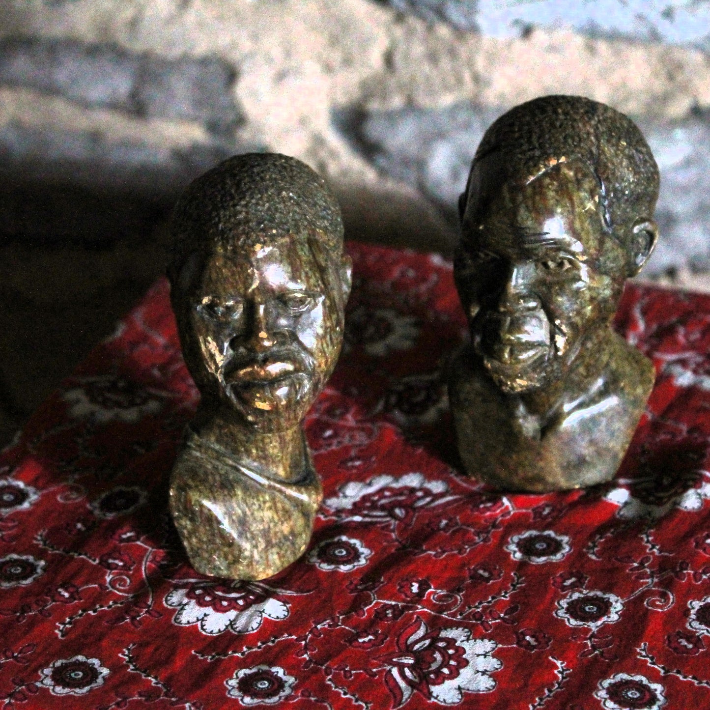 Two stone sculptures of faces on a red patterned fabric with a blurred background