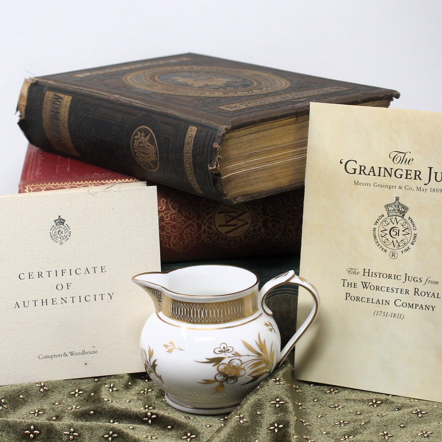 A porcelain creamer jug with gold floral decoration and a gold rim, displayed with its certificate of authenticity and an informational card on a green velvet surface, with antique books in the background.