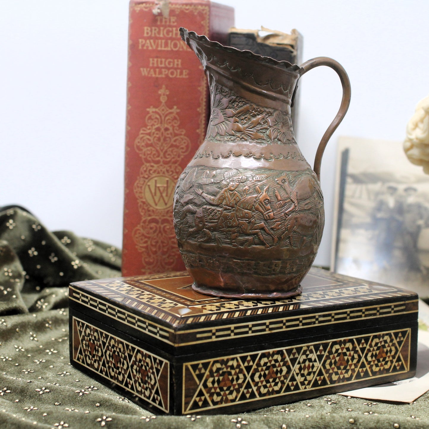An ornate decorative embossed copper pitcher with a curved handle and wide flared rim, displayed on top of a decorative black and gold patterned box. A red book titled 'The Brighton Pavilion' stands behind the pitcher, with soft-focus background objects visible.