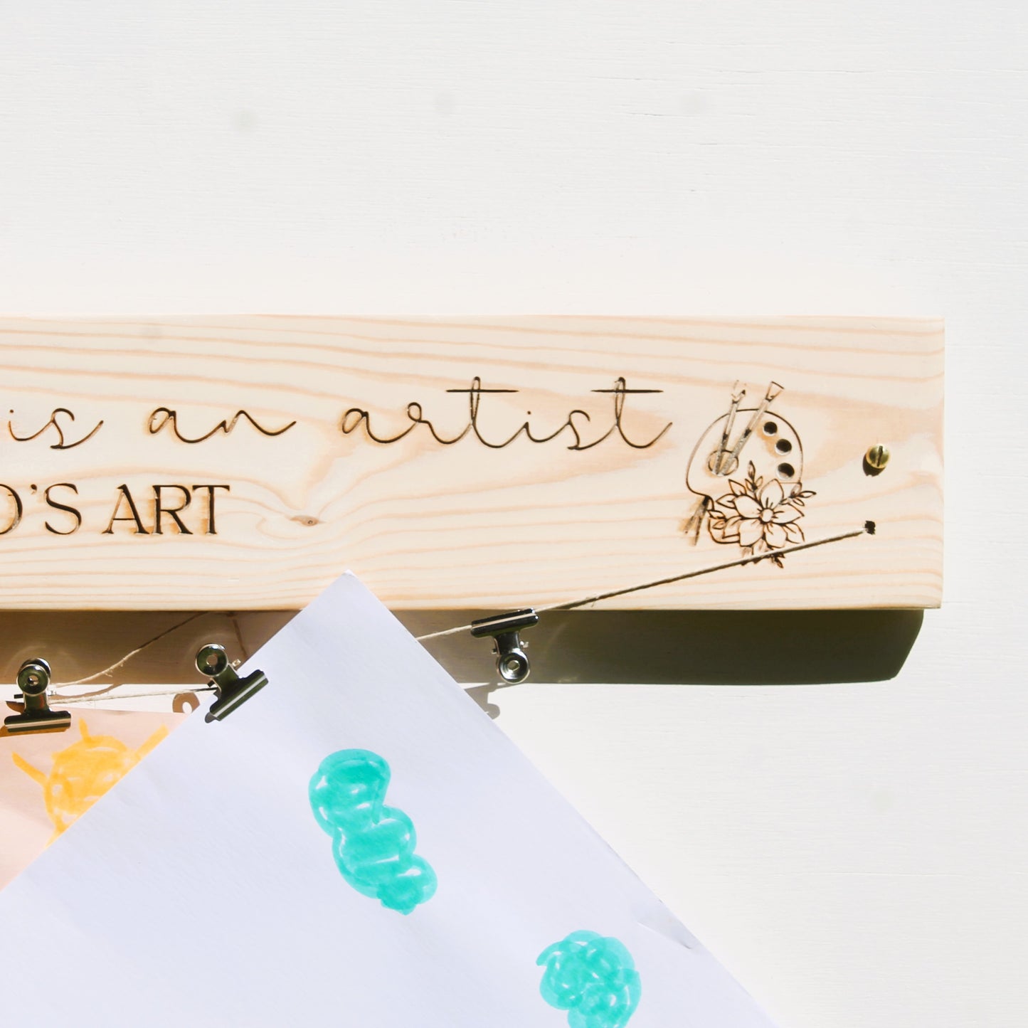Wooden art tool with 'is an artist O'S ART' engraving and a piece of paper with teal designs.