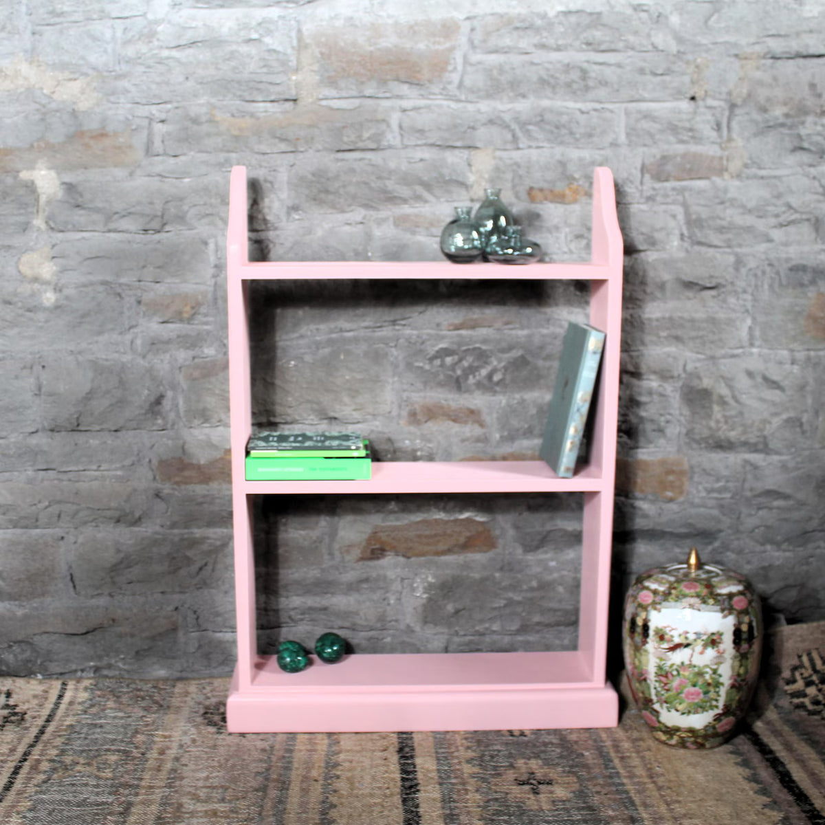 A pink wooden two-tier bookshelf with decorative items on it, set against a rustic gray brick wall background.