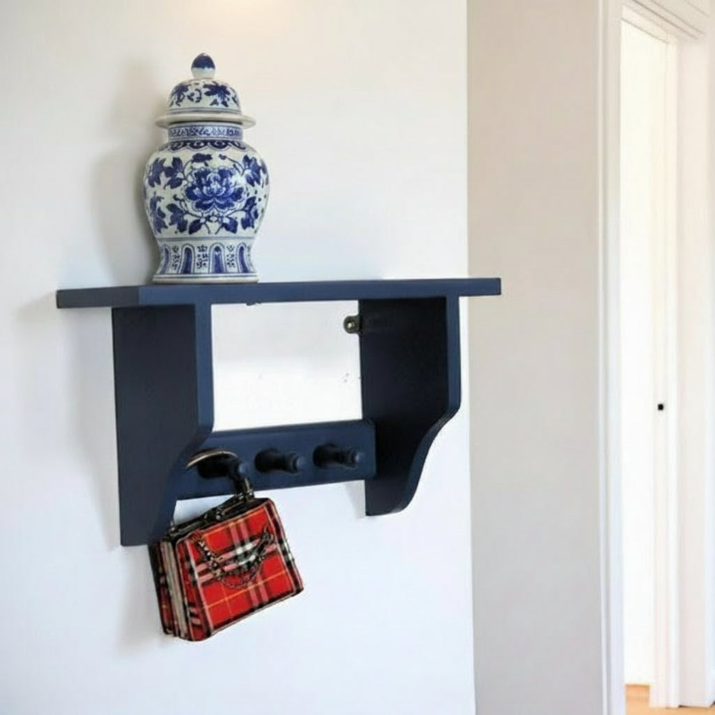 A navy blue wall-mounted shelf with three hanging hooks, styled in a home entryway. A chinese ginger jar sits on the shelf, and a red plaid handbag hangs from one of the hooks.