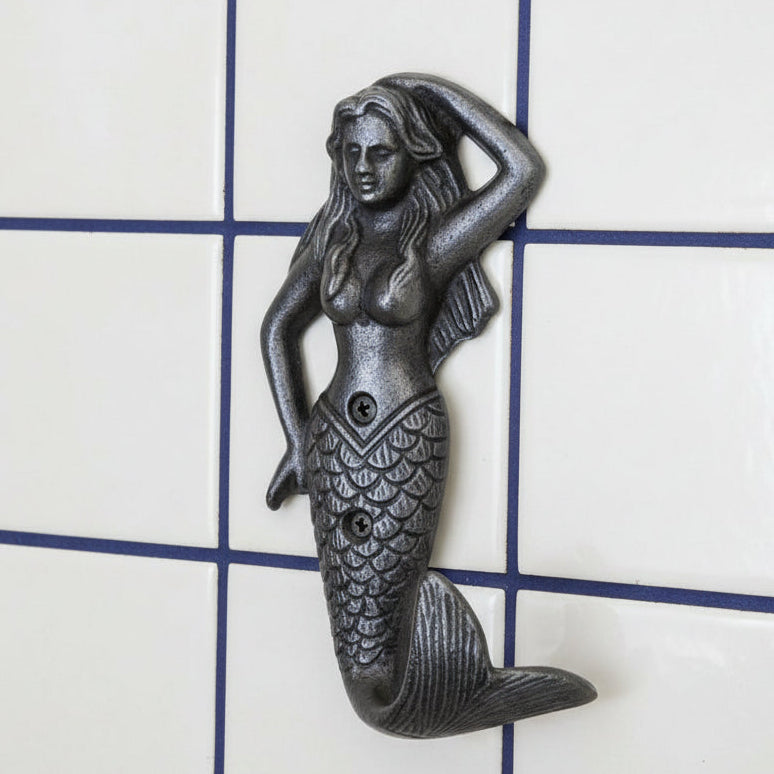 A cast iron mermaid wall hook with a textured, dark metallic finish, shown on a white tiled wall
