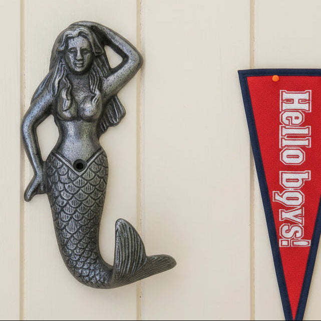 A cast iron mermaid wall hook with a textured, dark metallic finish, shown next to a wooden cladded wall with a red penant next to it