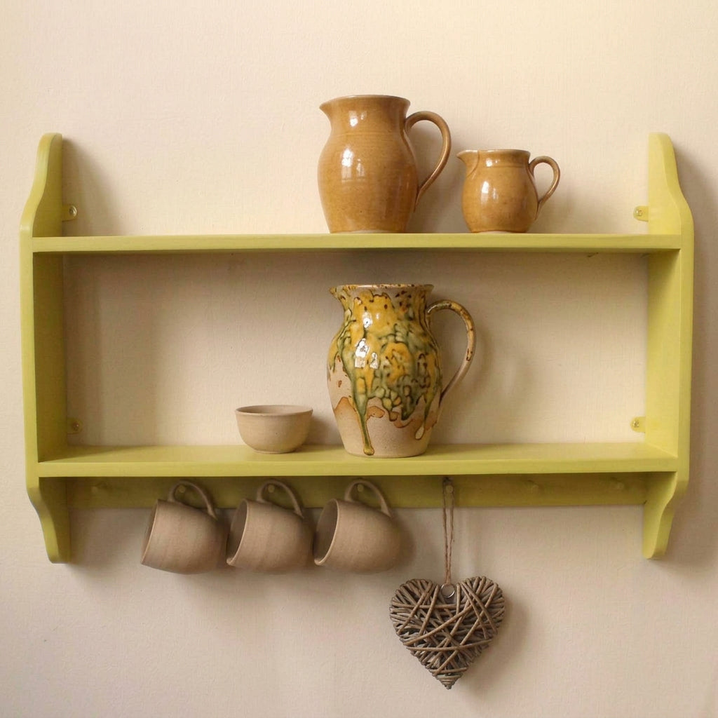 A light gray wooden two-tier wall shelf mounted on a beige wall, styled with white ceramic bowls, a red pitcher, striped mugs, dried lavender, and a woven heart decoration.