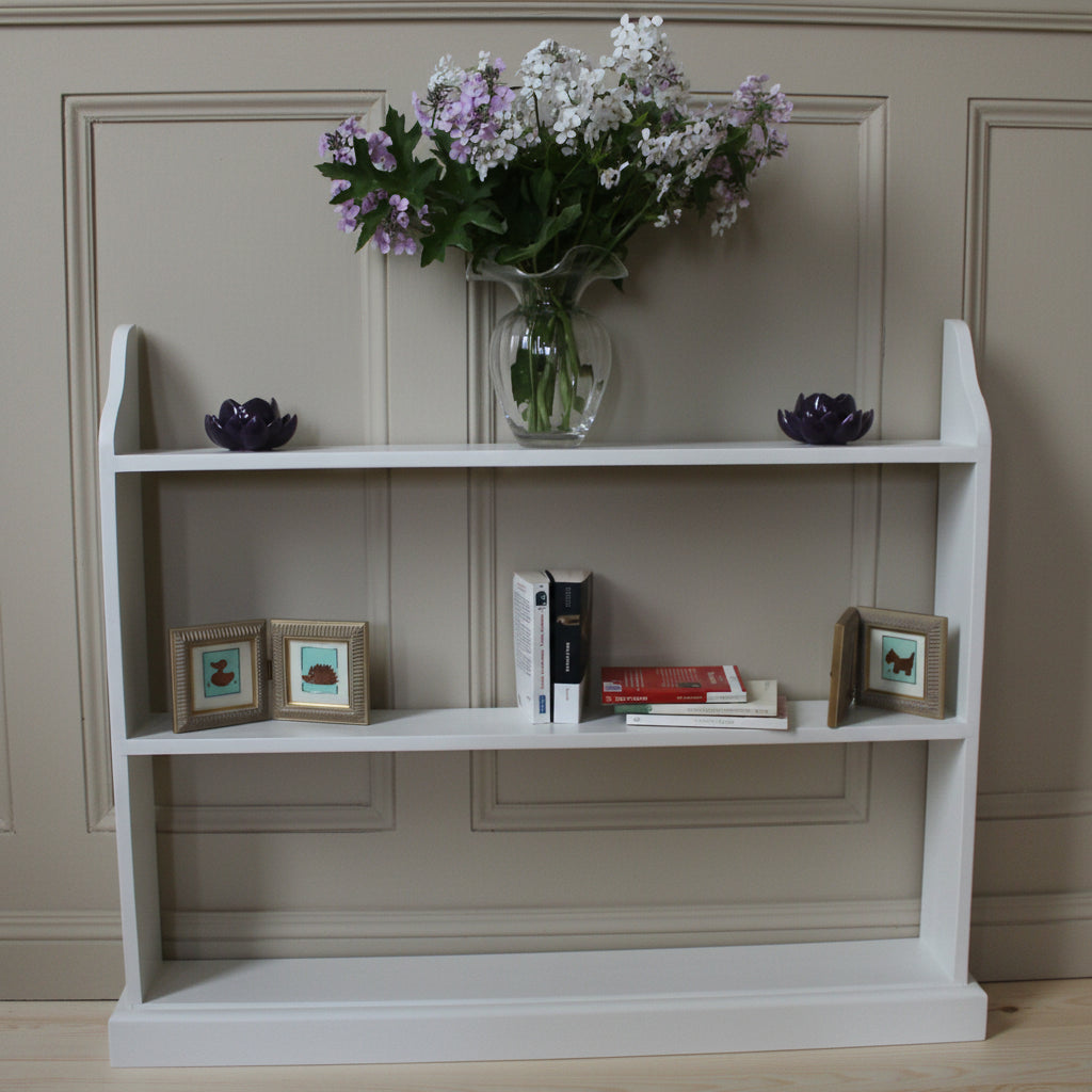 A white wooden open bookshelf with three tiers, styled with a glass vase of purple and white flowers, two dark purple lotus candles, two small framed pictures, and a stack of books, set against a plain off-white wall.