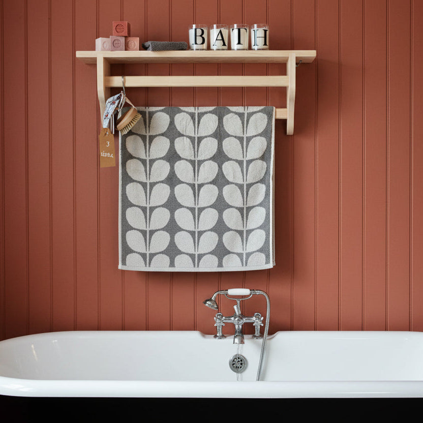 A bathroom scene with a freestanding white bathtub against a terracotta wood-paneled wall, a light wood shelf mounted above, and a gray-and-white leaf-patterned towel hanging from a hook.