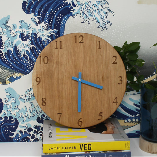 A round wooden clock with a natural oak finish and blue hands, styled on top of books against a blue wave-patterned wallpaper background.