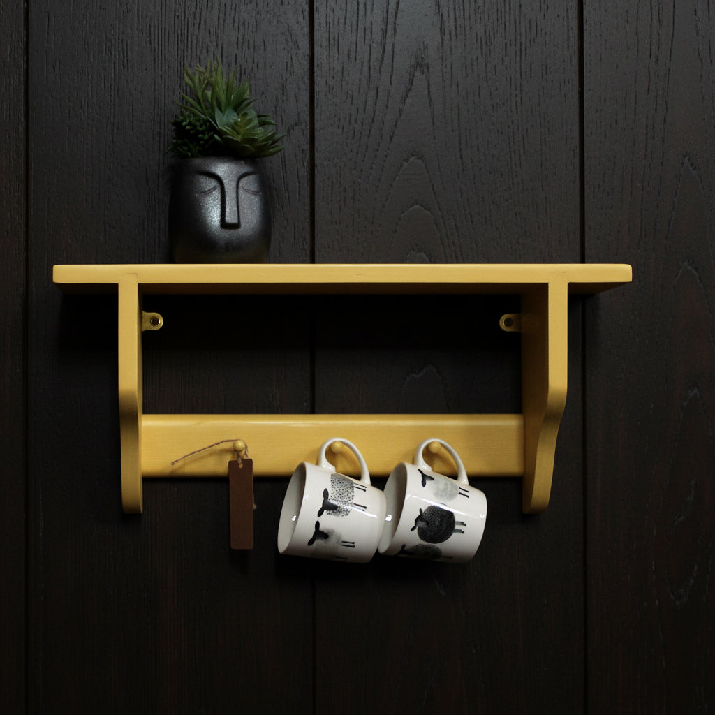 A mustard yellow wall-mounted shelf with a small potted succulent on top and two white ceramic mugs hanging underneath, set against a plain black wall.