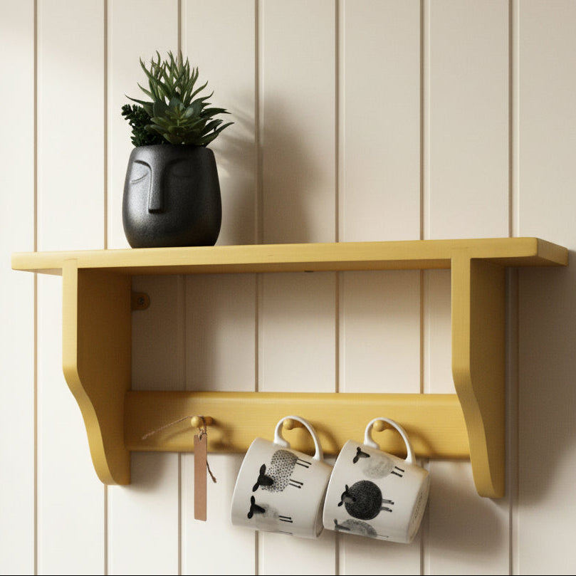 A mustard yellow wall-mounted shelf with a small potted succulent on top and two white ceramic mugs hanging underneath, set against a plain white wall.