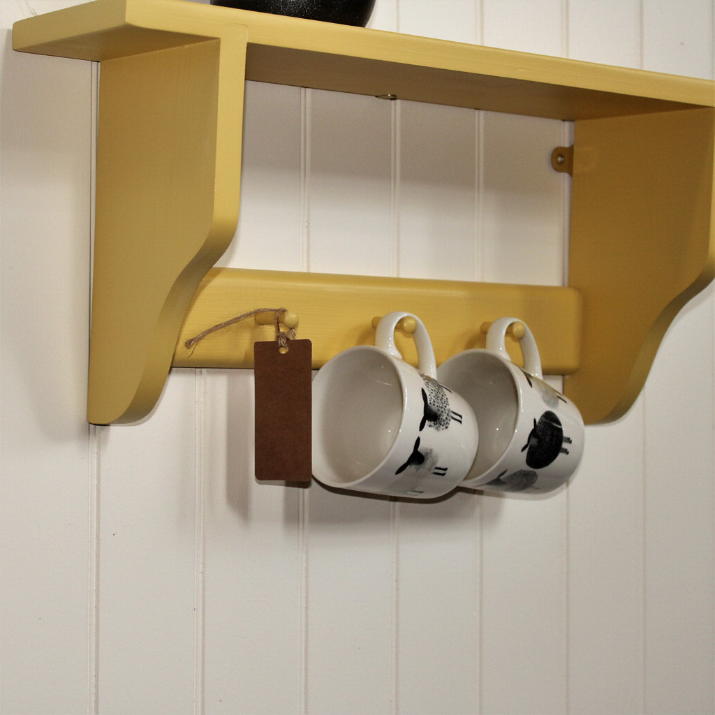 A mustard yellow wooden wall-mounted shelf with two hanging hooks holding two white ceramic mugs with black cow illustrations, against a white wall.