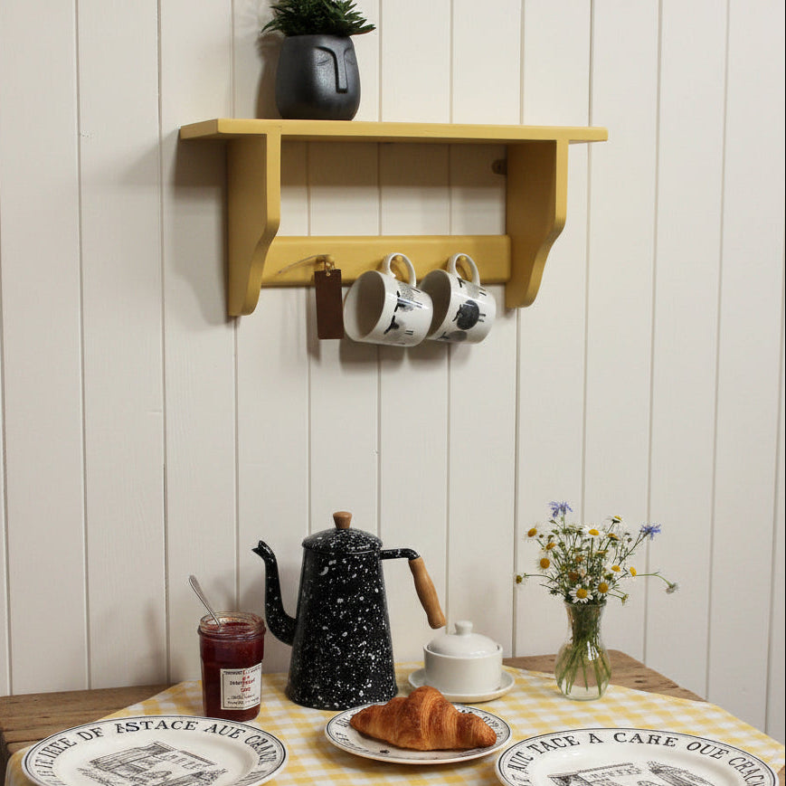 A mustard yellow wall-mounted shelf with a small potted plant on top and two white mugs hanging from the side hook, shown against a white wall.