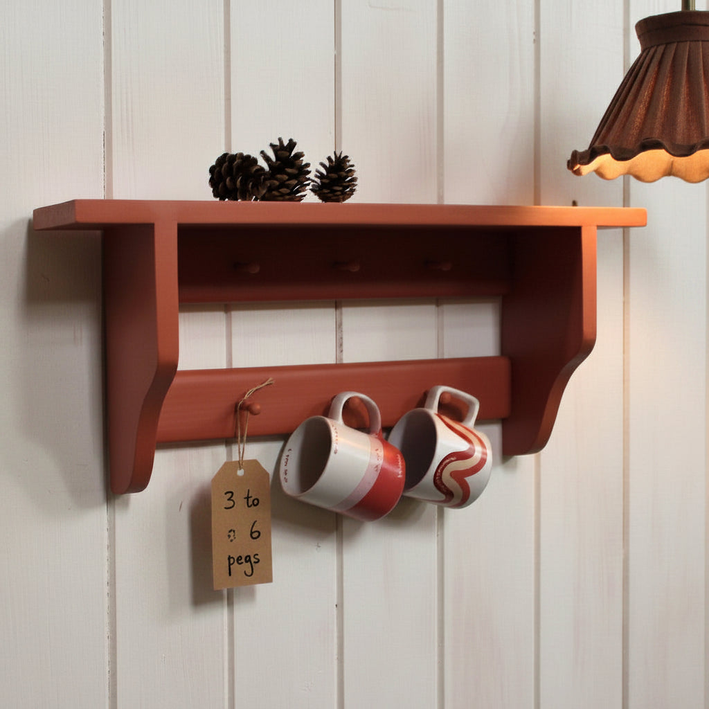 A reddish-brown wooden wall-mounted shelf with two pegs, shown against a white wall. Two white ceramic mugs with red swirl patterns hang from the pegs, and two pinecones rest on the top shelf.