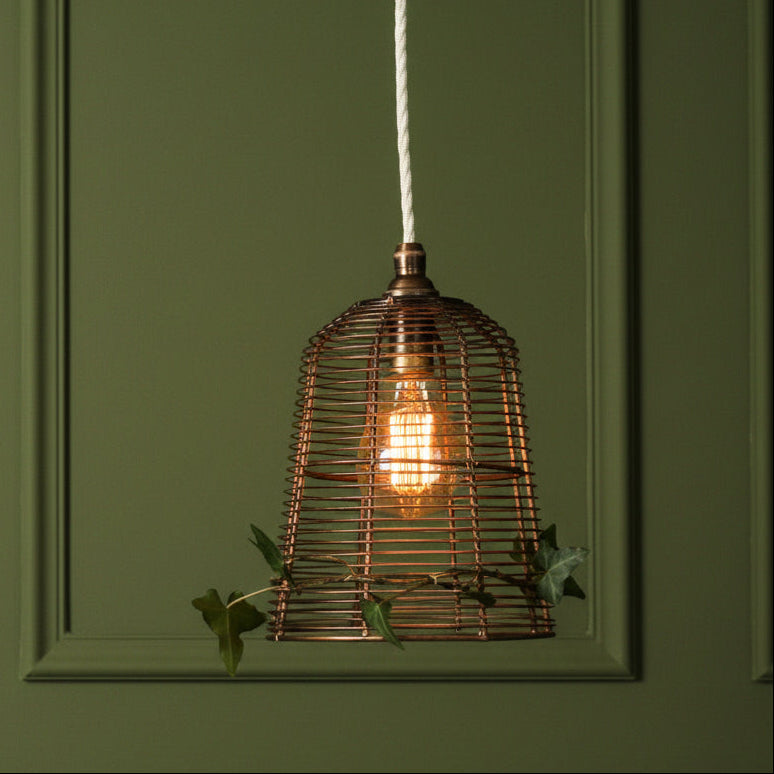 A copper wire cage pendant light hanging from a white cord against a dark green paneled wall, with a warm glowing bulb inside and small green leaves draped near the bottom.