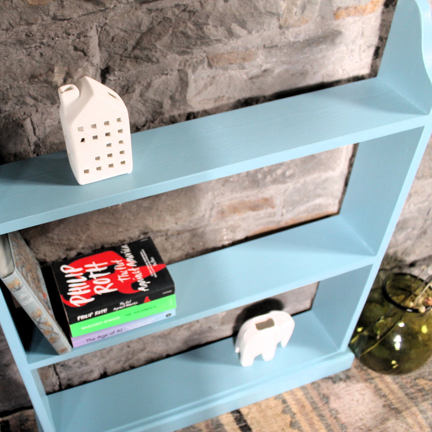 A light blue wooden bookshelf with three shelves, styled with a white ceramic house-shaped candle holder, a book, and a small white elephant figurine, set against a rustic stone wall.