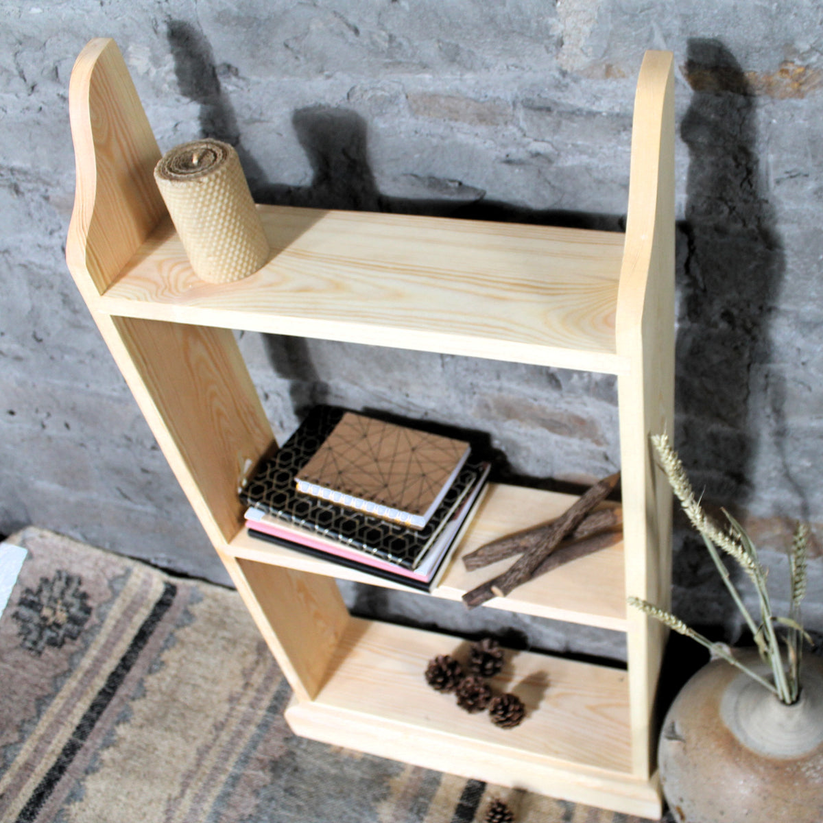 A light natural wood three-tier shelf with curved side supports, styled with books, a rolled notebook, twigs, pinecones, and dried wheat in a ceramic pot, set against a gray brick wall.
