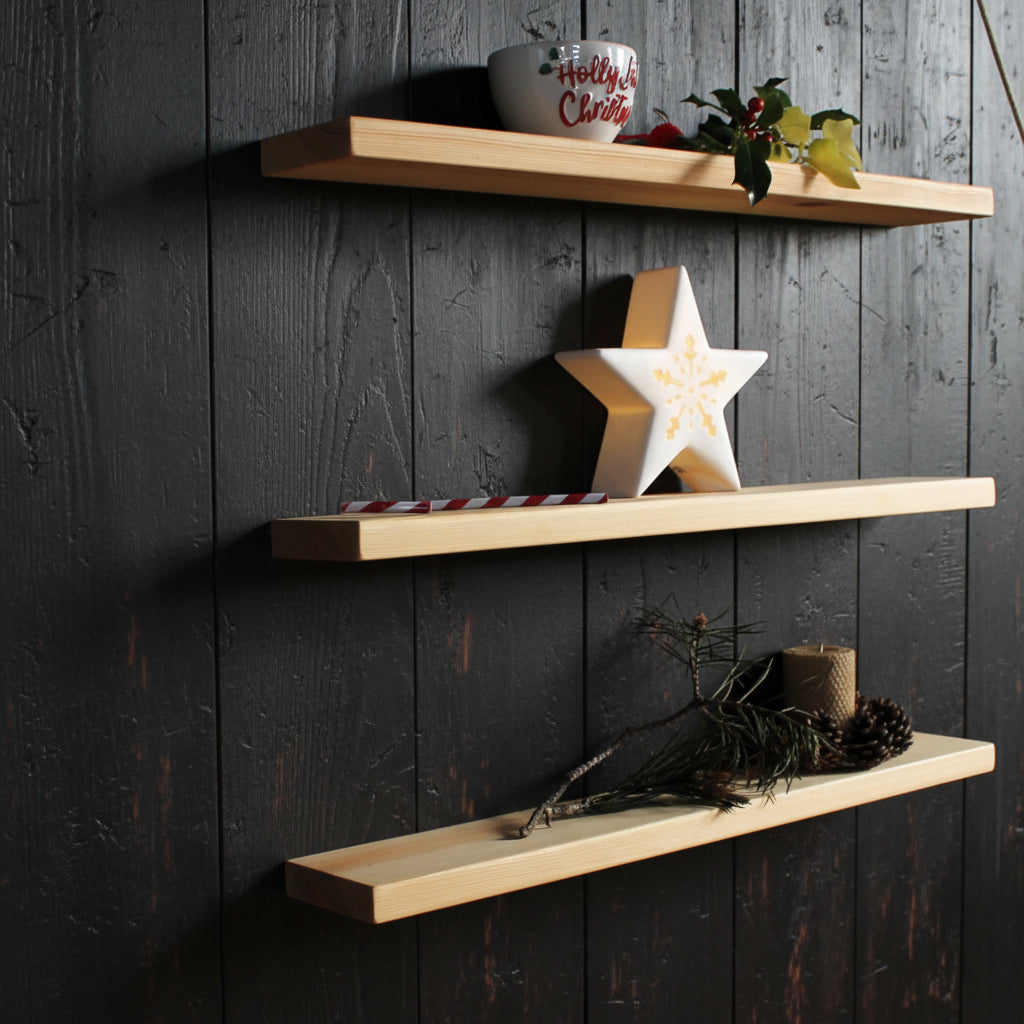 Three light natural wood floating shelves mounted on a dark wall, styled with holiday decorations including a white mug, a wooden star, a candy cane, pine branches, and a candle.
