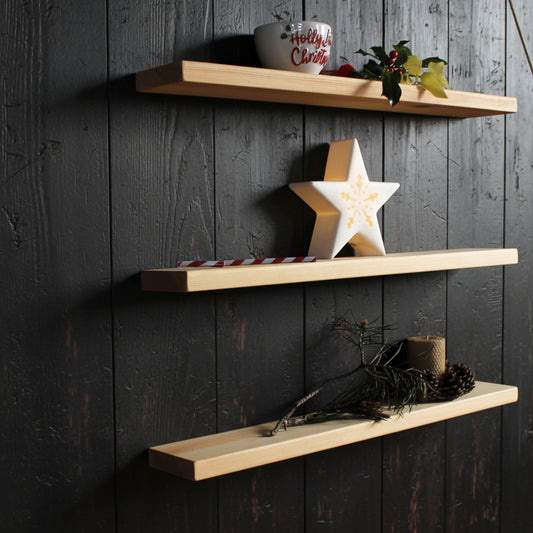 Three light natural wood floating shelves mounted on a dark wall, styled with holiday decorations including a white mug, a wooden star, a candy cane, pine branches, and a candle.