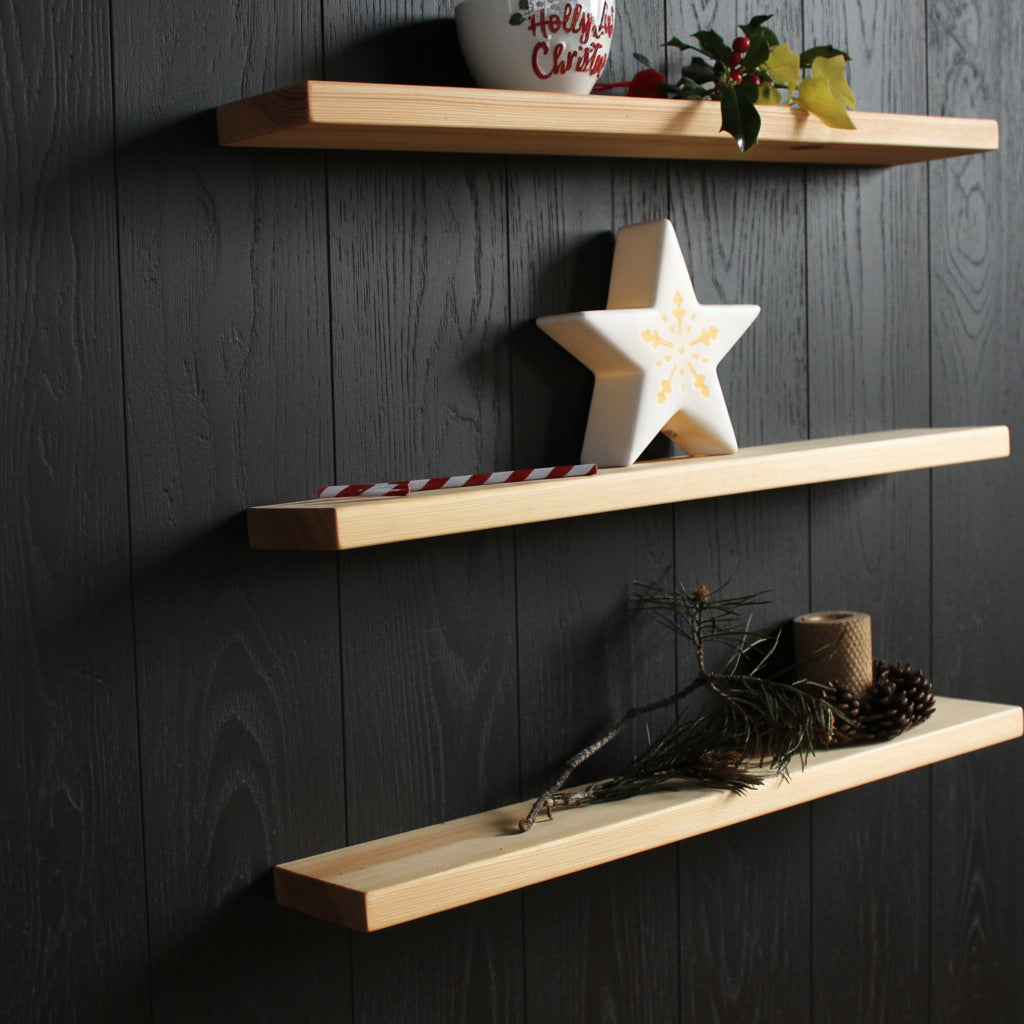 Three light natural wood floating shelves mounted on a black wall, styled with holiday decorations including a white star ornament, a small potted plant, and pinecones.