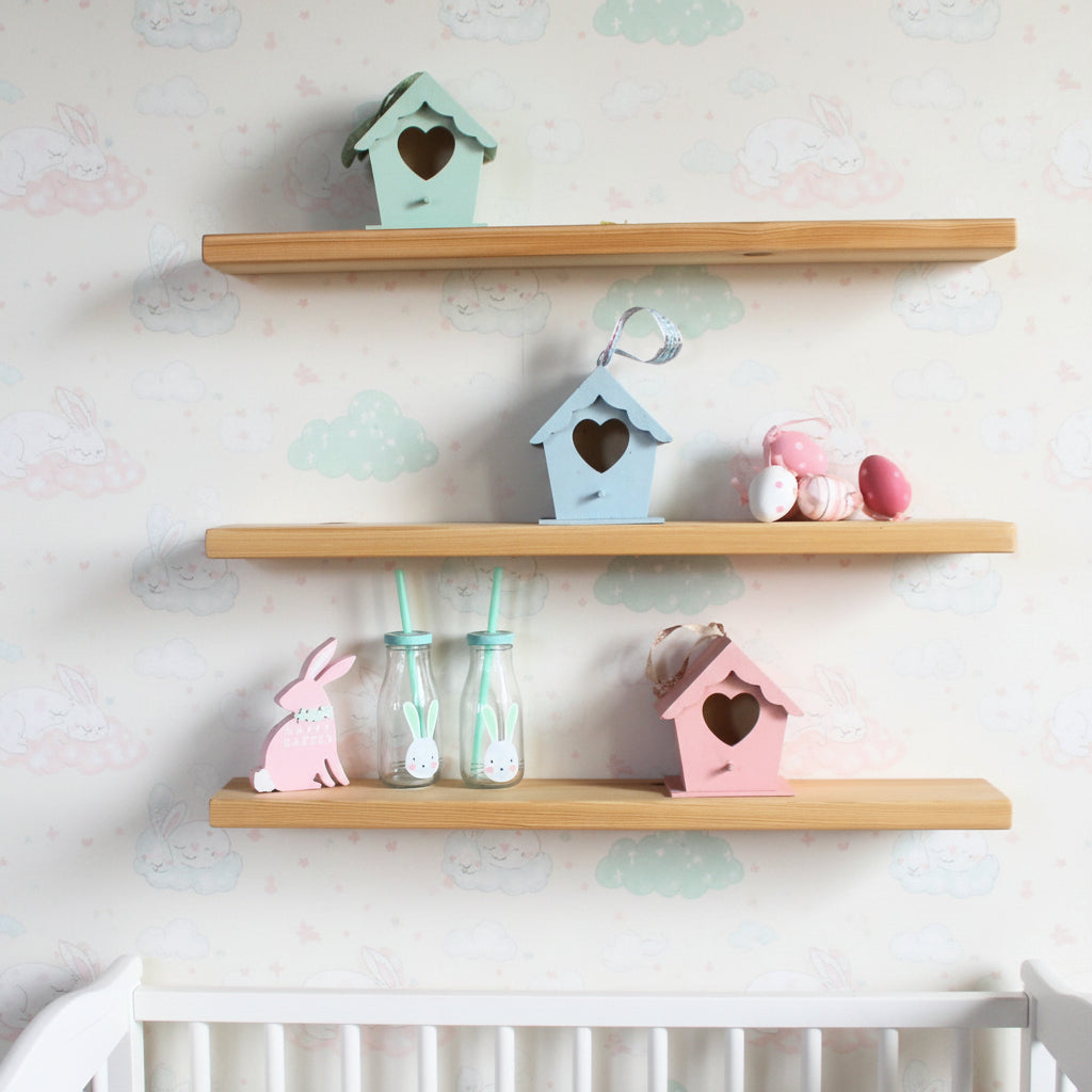 Three light wood floating shelves mounted on a white wall, styled with pastel decorative items including small birdhouses in mint, light blue, and pink, a pink bunny cutout, two glass bottles with mint straws, and a small plush toy.