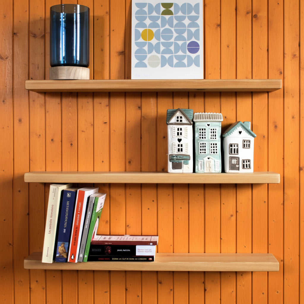 Three light natural wood floating shelves mounted on an orange wall, styled with a blue glass vase, a geometric art print, small decorative house figurines, and a few books.