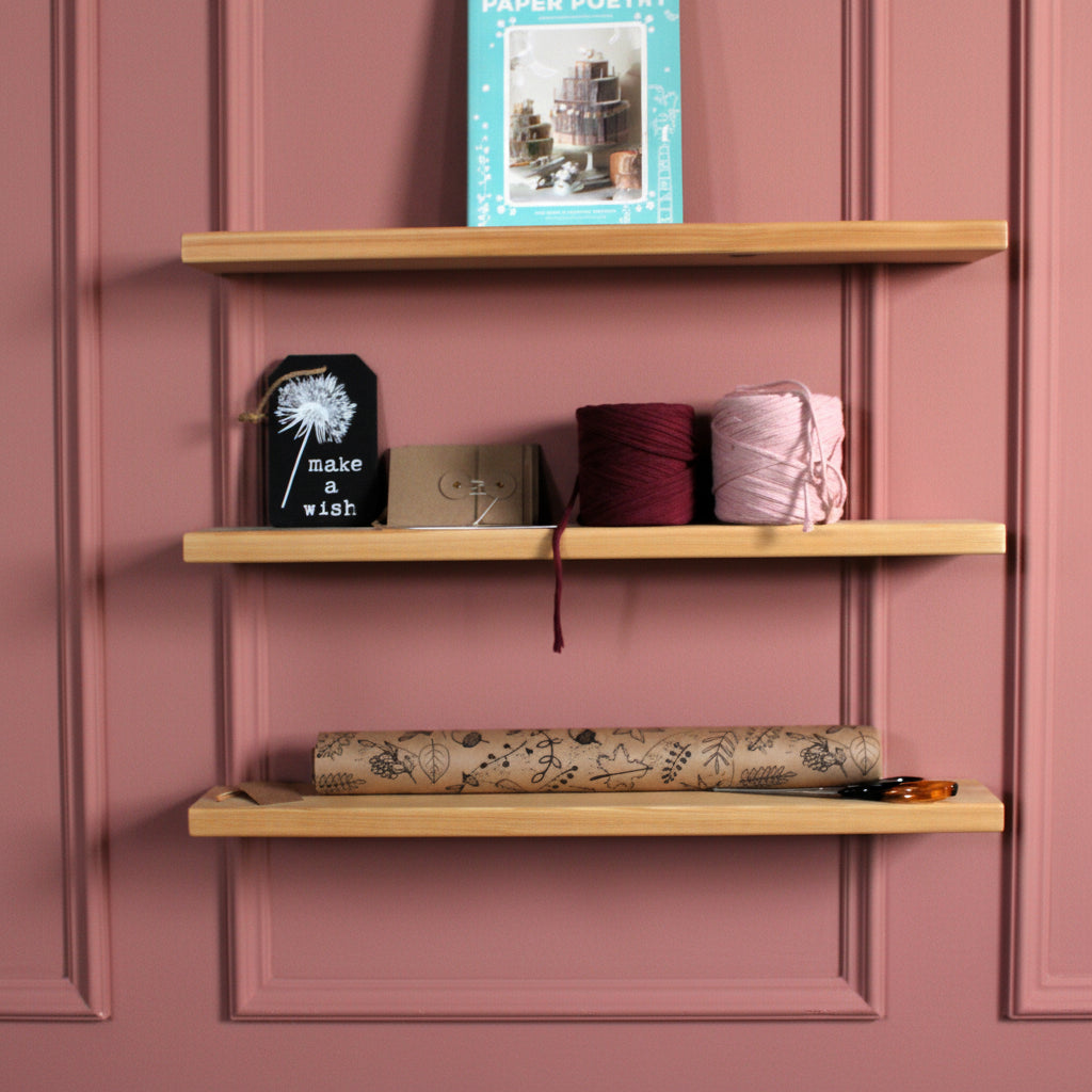 Three light natural wood floating shelves mounted on a pink wall, styled with a book, a small black sign, yarn balls, and a patterned fabric roll.