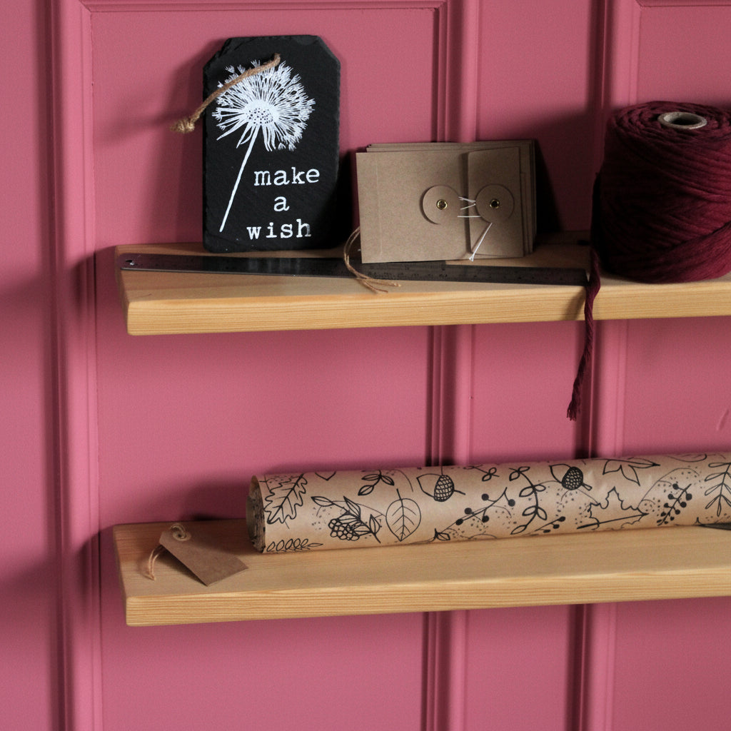 Two light natural wood floating shelves mounted on a pink wall, styled with a black chalkboard tag reading 'make a wish', a small brown envelope, a spool of dark red thread, and a rolled piece of brown patterned paper.