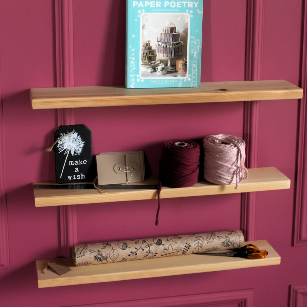 Three light natural wood floating shelves mounted on a pink wall, styled with books, yarn balls, a small sign, and a rolled piece of patterned fabric.
