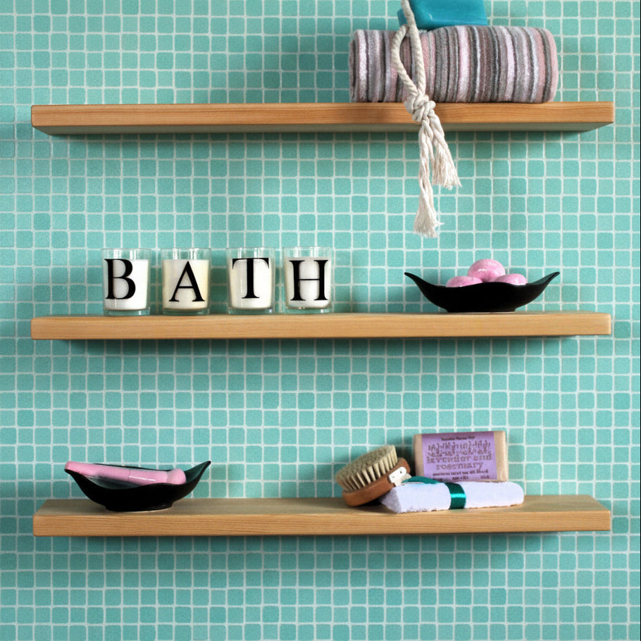 Three light natural wood floating shelves mounted on a turquoise wall, styled with bath and beauty items including a striped towel, candles spelling B-A-T-H, a black soap dish, and a brush.