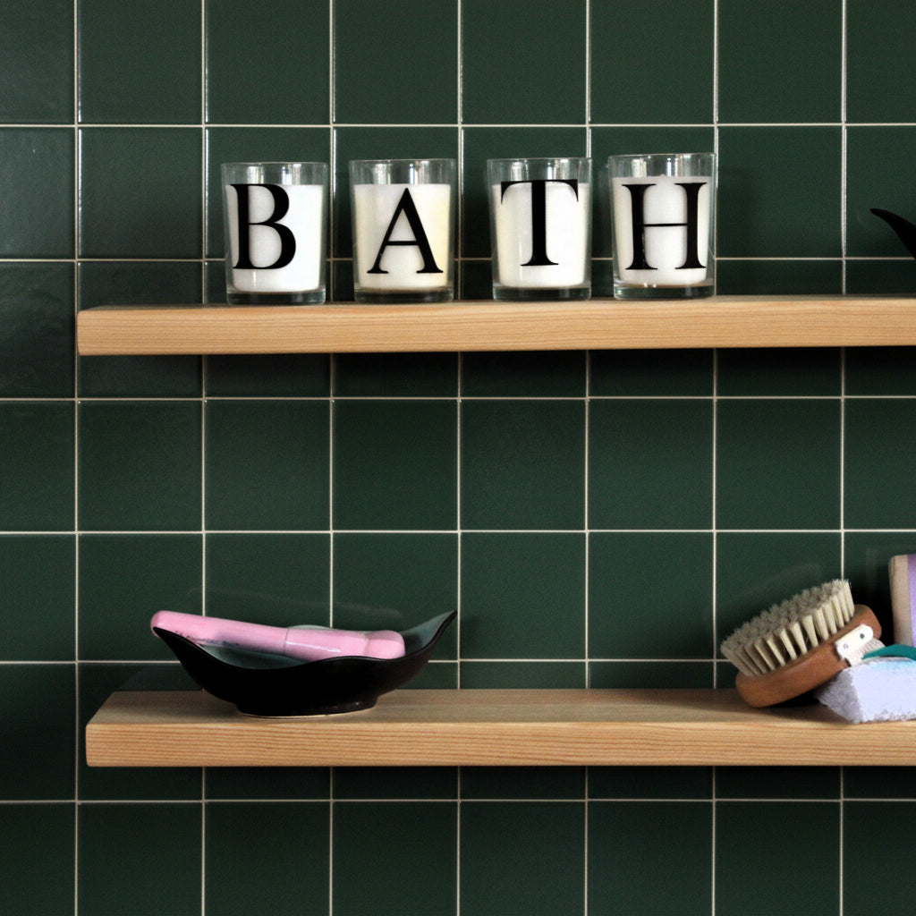 Four clear glass candles arranged on a light wood floating shelf, each with a white wax insert and a large black letter, spelling B-A-T-H. The shelf is mounted on a green wall above another shelf holding a black dish with a pink lid and a brush.