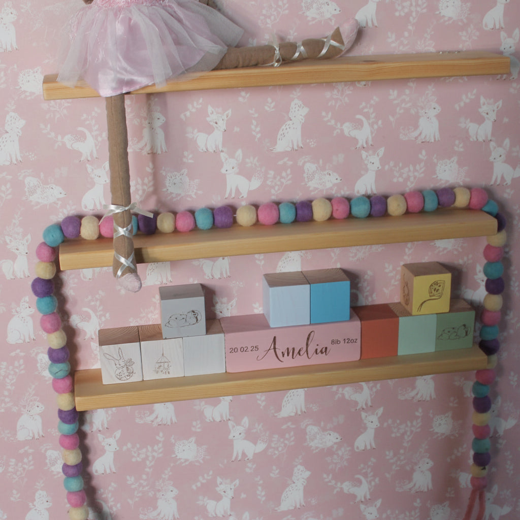 A wooden children's shelf display with pastel pom-pom garlands, wooden blocks, and a ballerina doll on apink wall.
