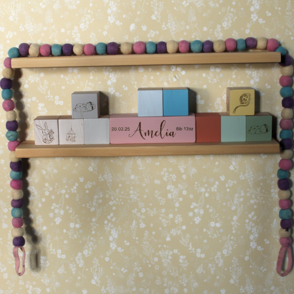 A wooden two-shelf wall display decorated with colorful felt beads and holding custom wooden blocks, including one personalized with the name 'Amelia' and another with a bunny illustration.