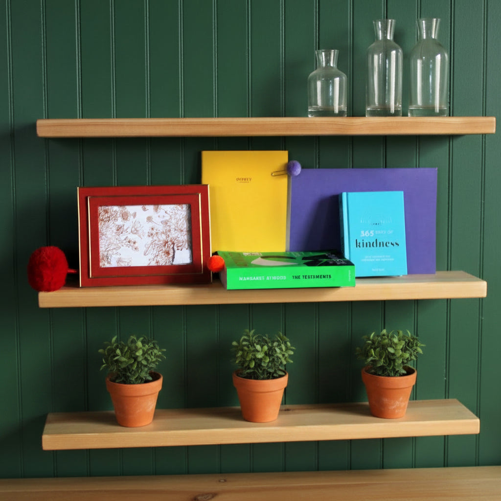 Three light natural wood floating shelves mounted on a white wall, styled with colorful books, a red picture frame, and small decorative pom-poms.