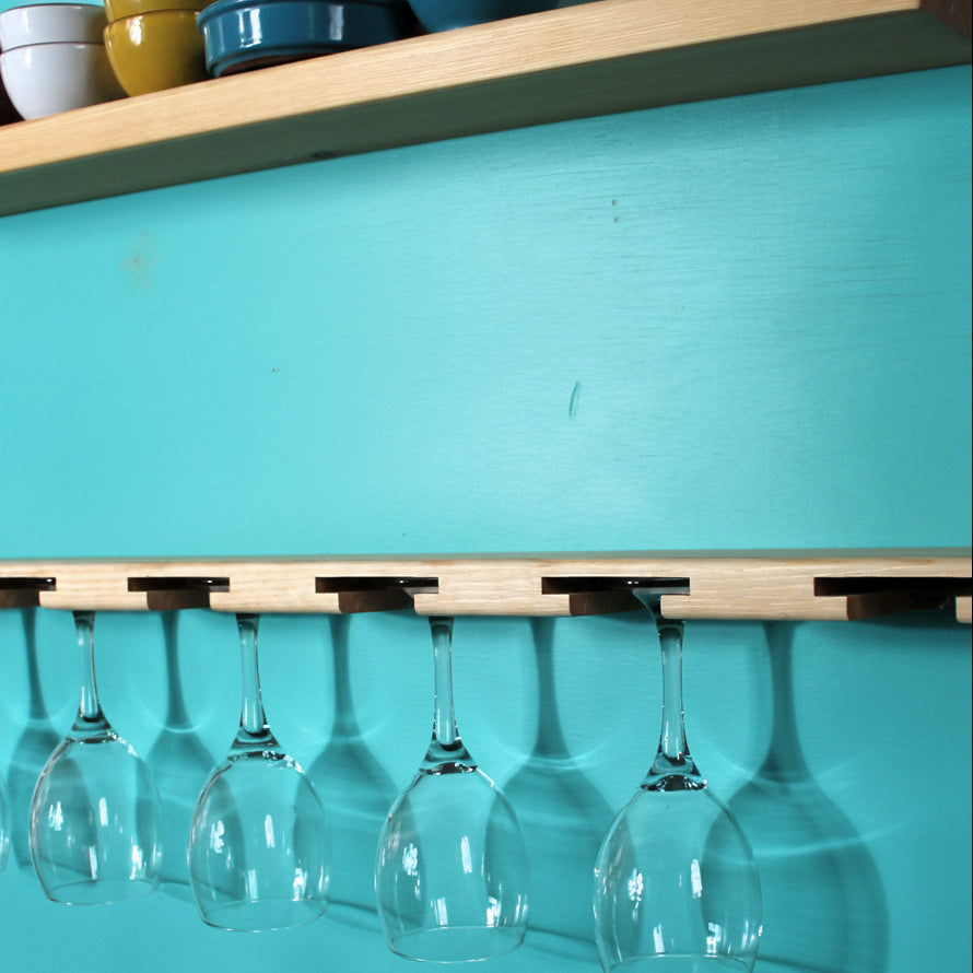 A light natural wood wall-mounted glass holder with six slots, shown holding six clear wine glasses upside down against a turquoise wall. A matching wooden shelf with colorful bowls is visible above.