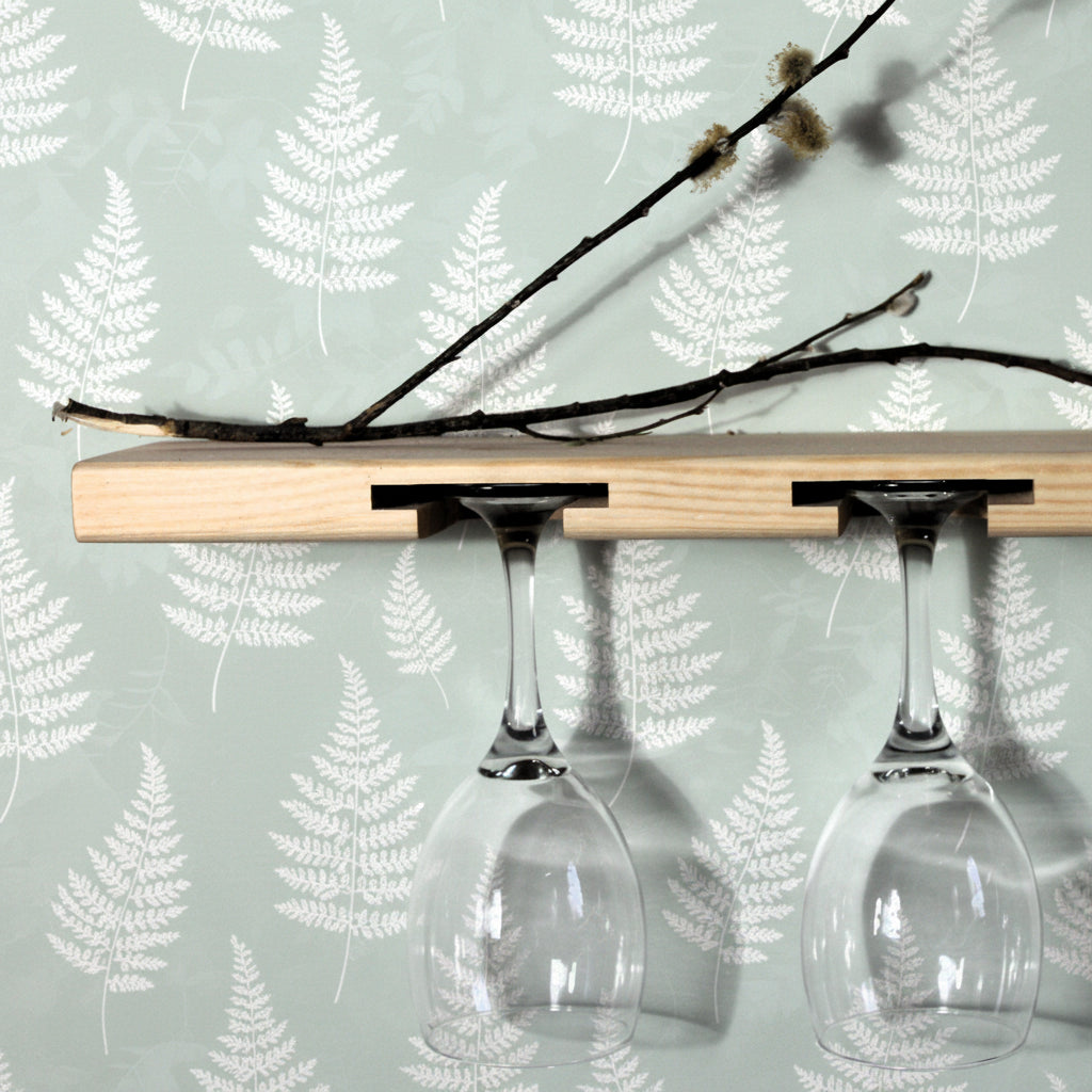 A light natural wood wall-mounted wine glass holder with two cutouts, shown holding two clear wine glasses upside down against a plain white wall.