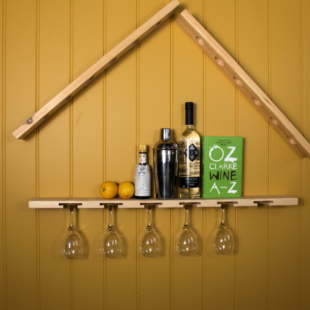 A wooden house-shaped wall shelf mounted on a white wall, holding five hanging wine glasses, several liquor bottles, two lemons, and a green book.
