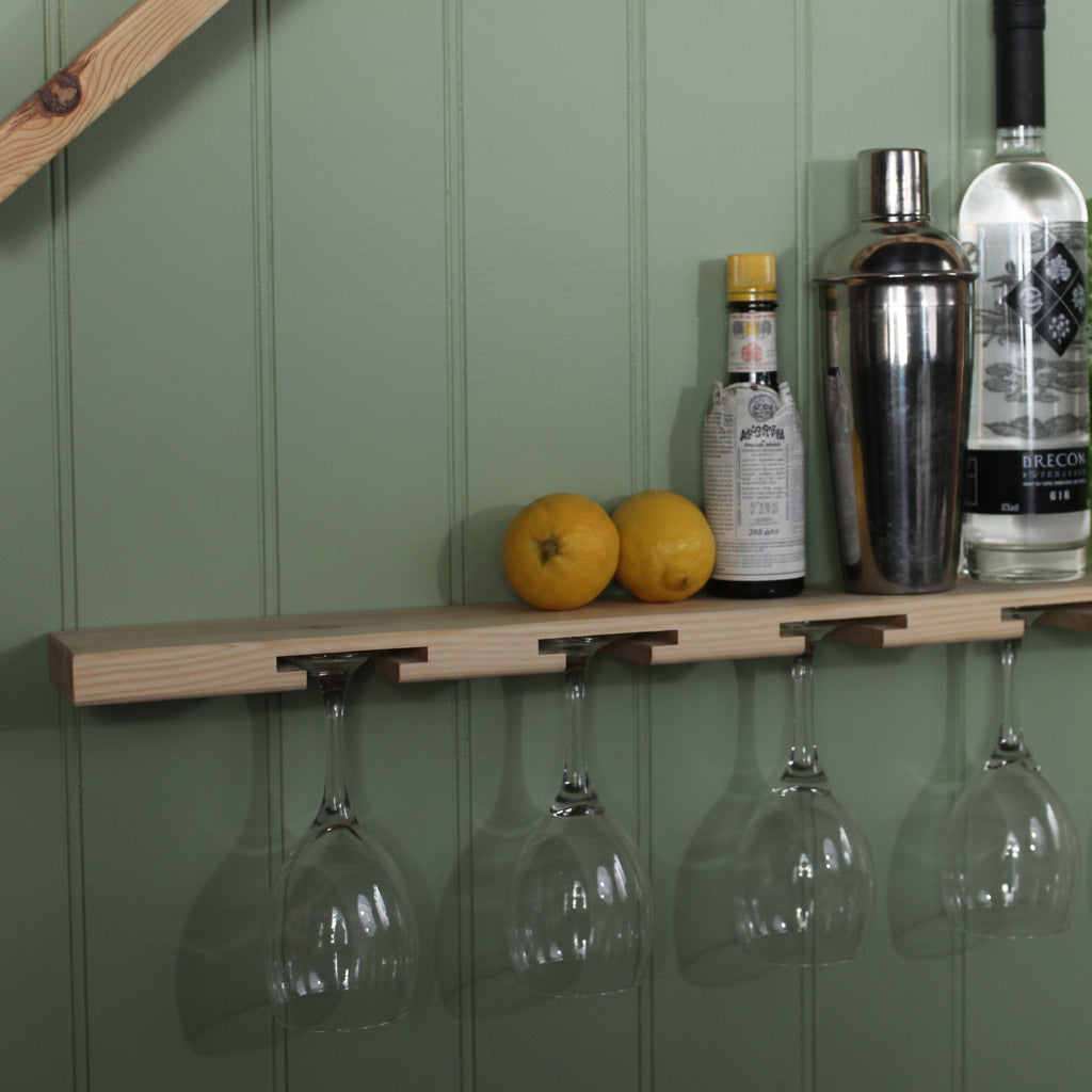 A light natural wood wall-mounted shelf holding wine glasses hanging from cutouts, with bottles, lemons, and a green book on top, against a white wall.