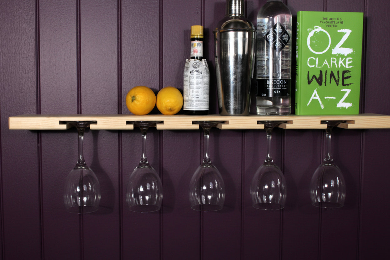 A light wood wall-mounted wine glass rack with five hooks holding five clear wine glasses upside down. On the shelf above, two lemons, a liquor bottle, a metal shaker, and a green book titled 'Oz Clarke Wine A-Z' are arranged against a plain white wall.