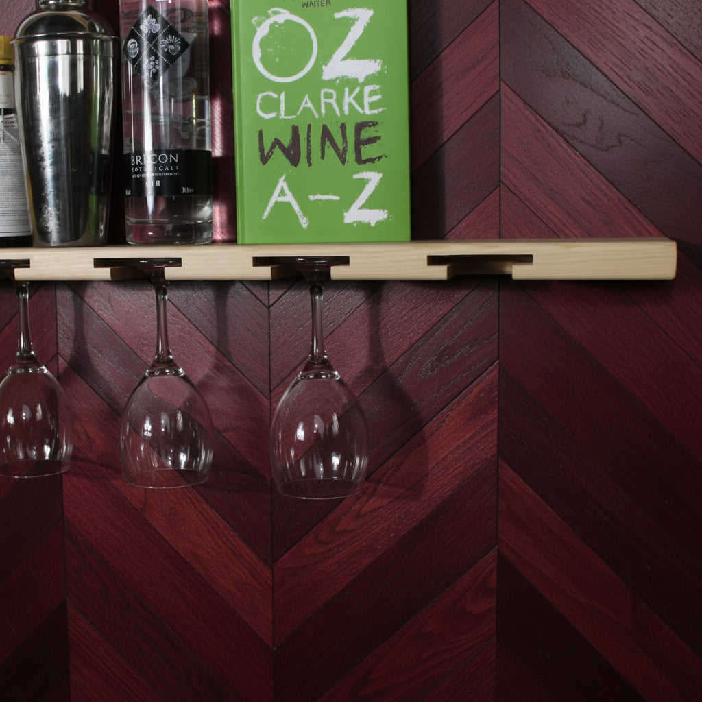 A light wood wall-mounted wine glass rack with six hooks holding six wine glasses upside down, shown against a white wall with bottles and lemons on top.