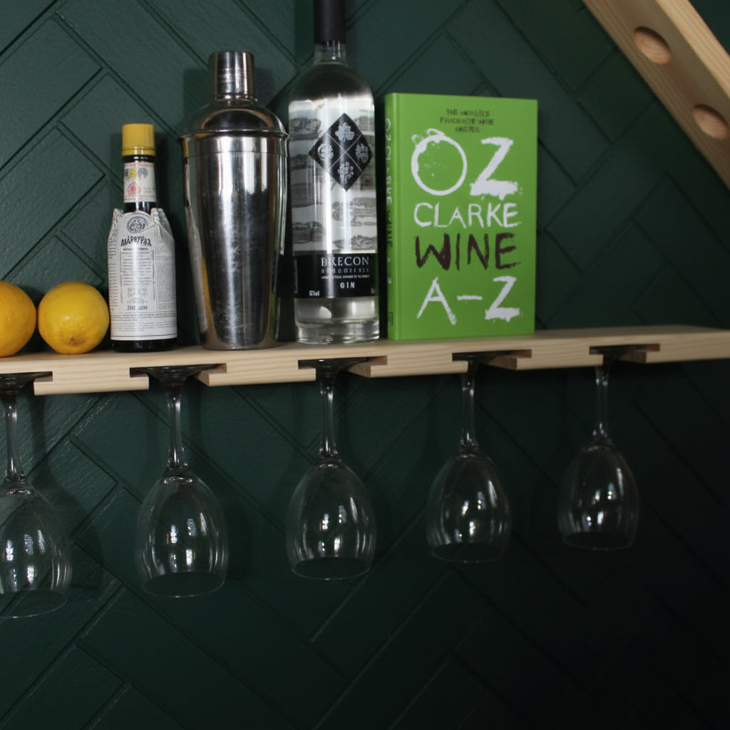 A wooden wall-mounted shelf holding wine glasses hanging below, with bottles and a green book on top against a white wall.