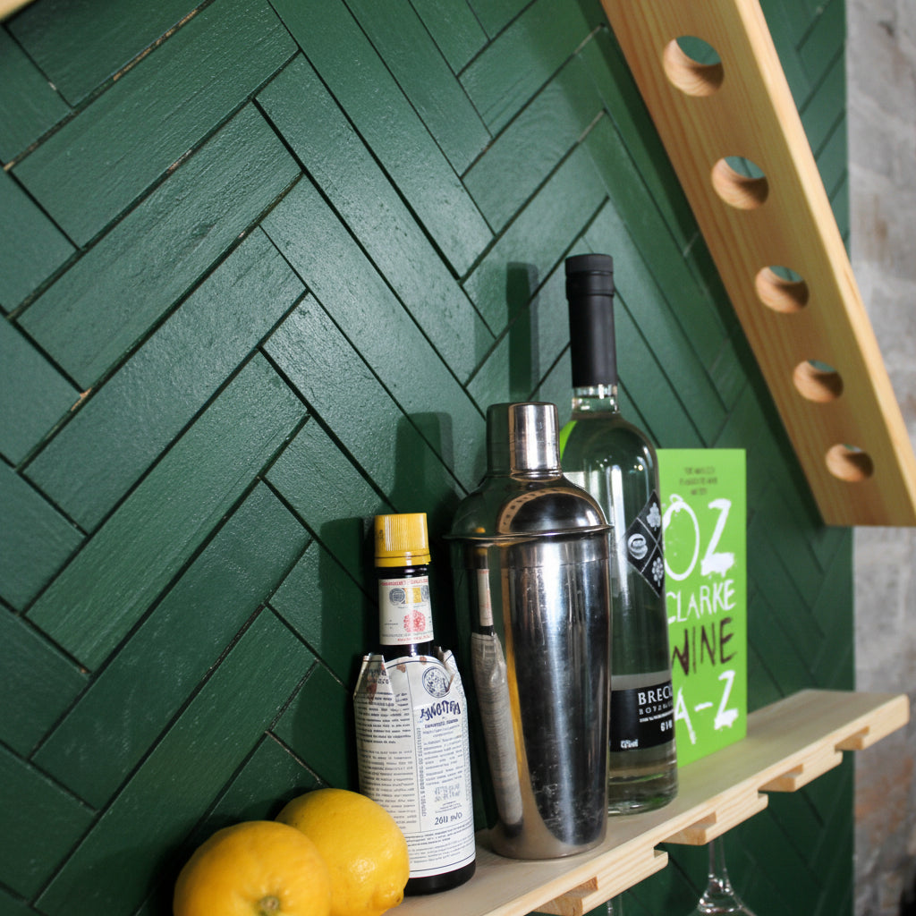 A wooden shelf holding a stainless steel cocktail shaker, a green bottle, a small bottle with a yellow cap, a green book, and two oranges, against a white wall with a wooden slat visible in the background.