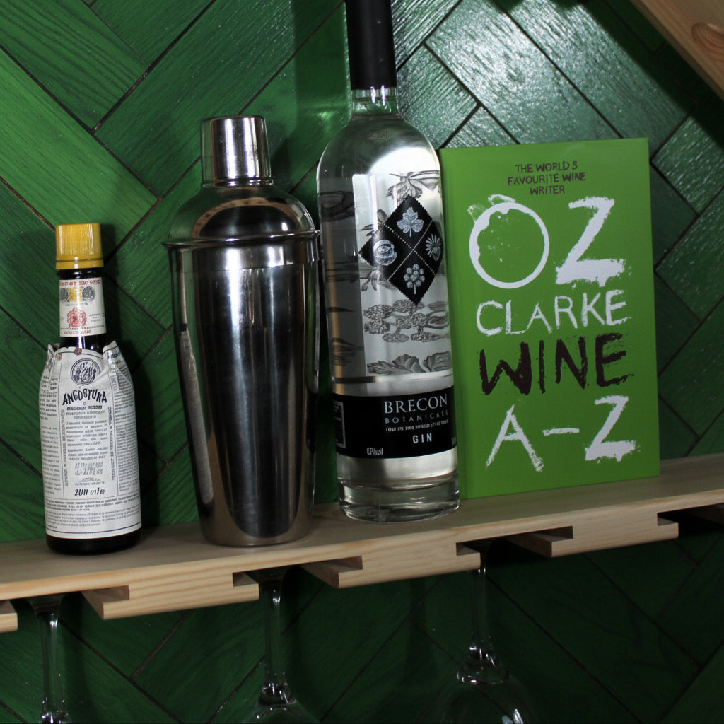 A wall-mounted wooden wine rack with diagonal support beams and a lower shelf, styled with wine bottles, lemons, a cocktail shaker, and a green book against a white wall.