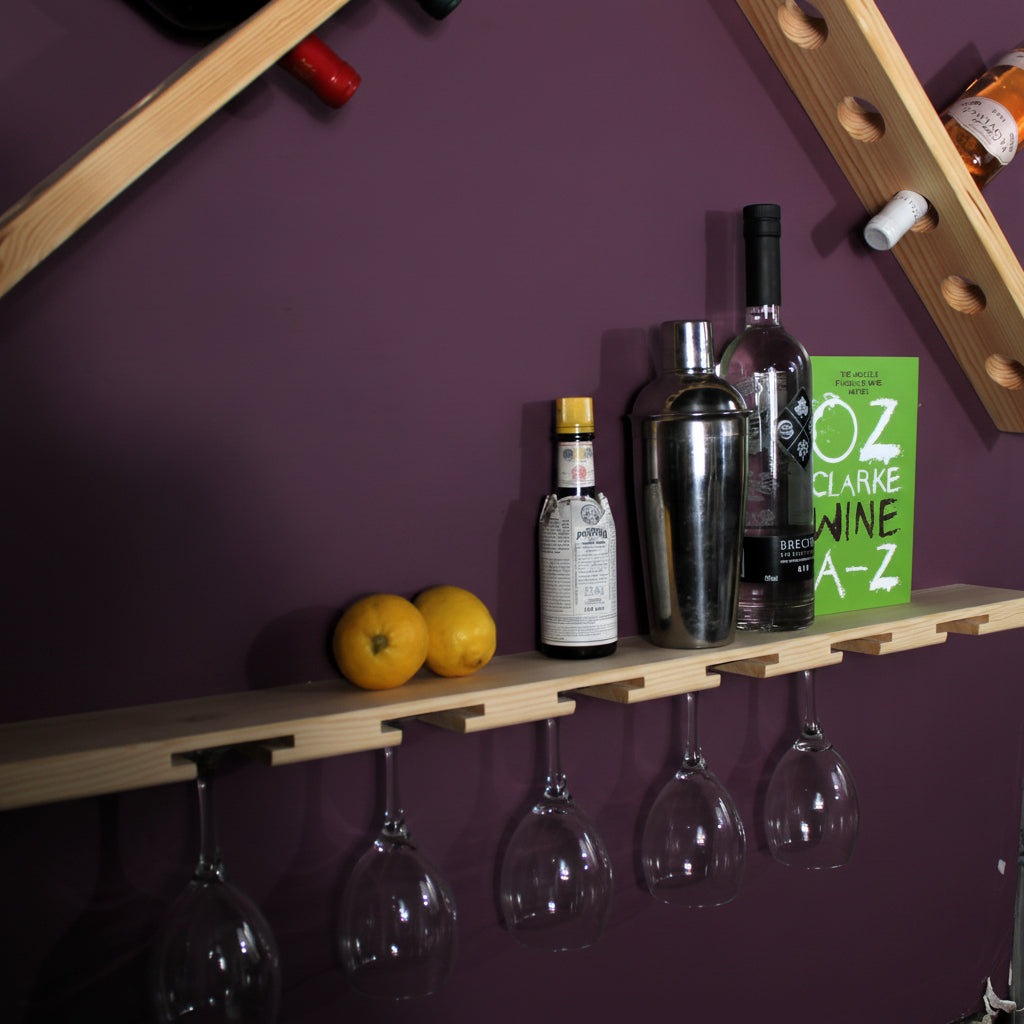 A wooden wall-mounted bar shelf holding wine glasses, bottles, two oranges, and a green book against a white wall.