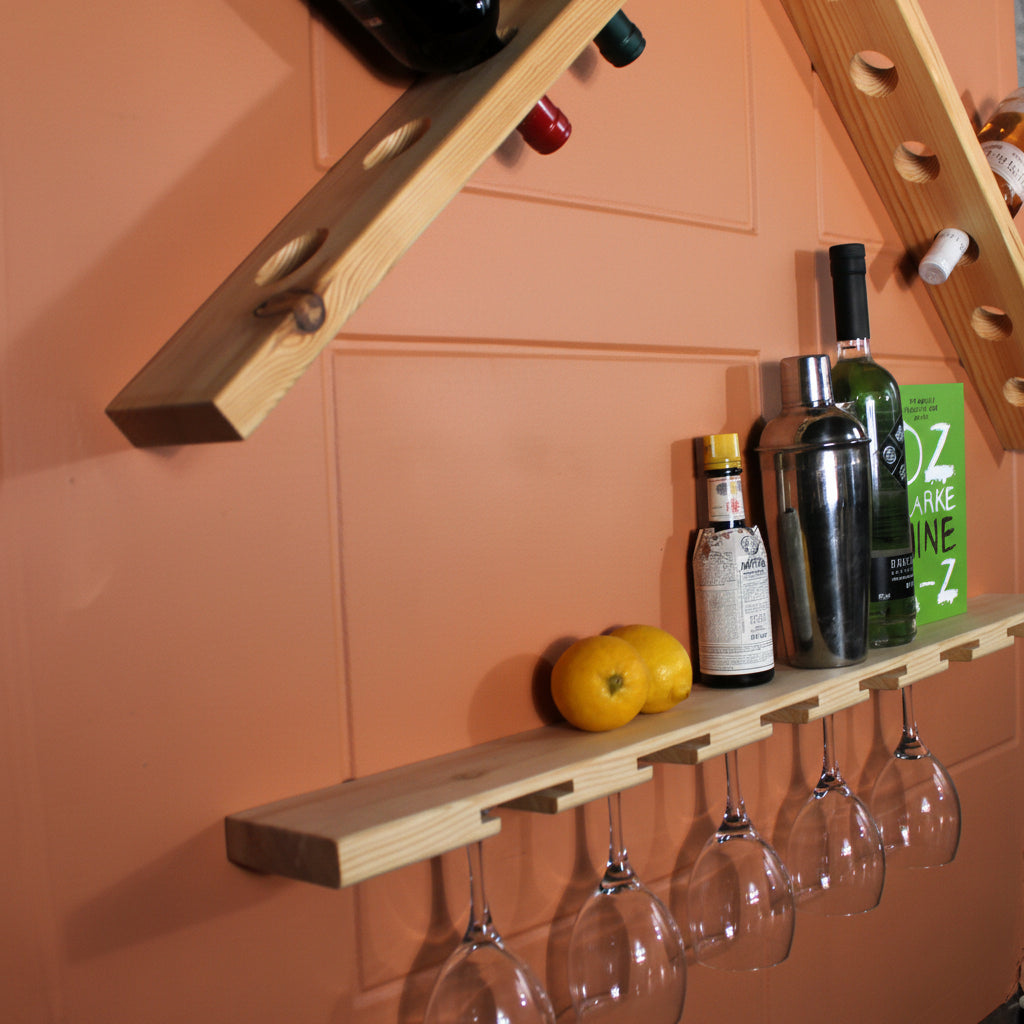 A light natural wood wall-mounted wine rack with multiple angled shelves, shown holding several wine bottles and several clear wine glasses hanging from the lower shelf. The background is a plain white wall.