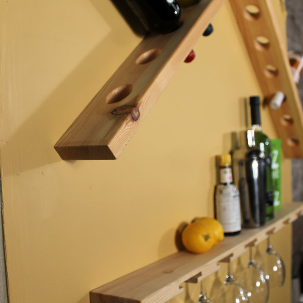 A light natural wood wall-mounted wine rack with angled beams, holding a dark bottle at the top and several wine glasses hanging from the lower shelf. A yellow fruit and a few bottles sit on the shelf against a white wall.