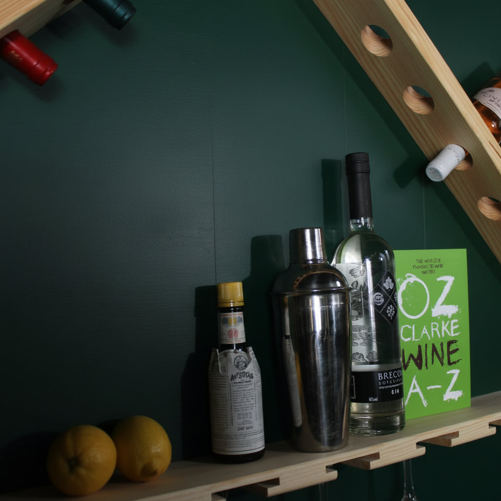 A wooden wall-mounted wine bottle rack with diagonal shelves holding several wine bottles, alongside a cocktail shaker, a small bottle, oranges, and a green book on a white wall.