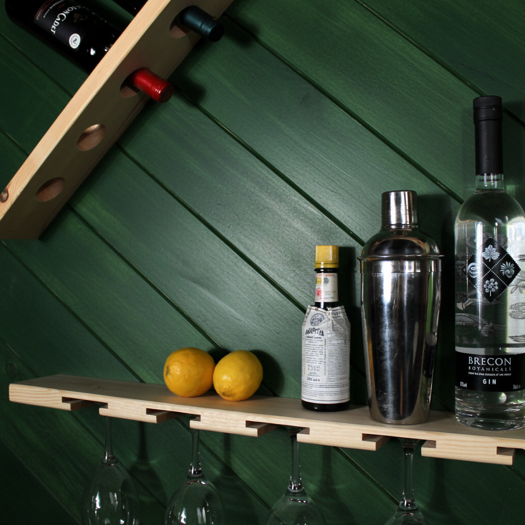 A light natural wood wall-mounted bar shelf with integrated bottle holders, wine glass hooks, and a shaker mount, styled with two lemons, a metal cocktail shaker, a bottle of Brecon Non-Alcoholic Gin, and wine glasses hanging below.