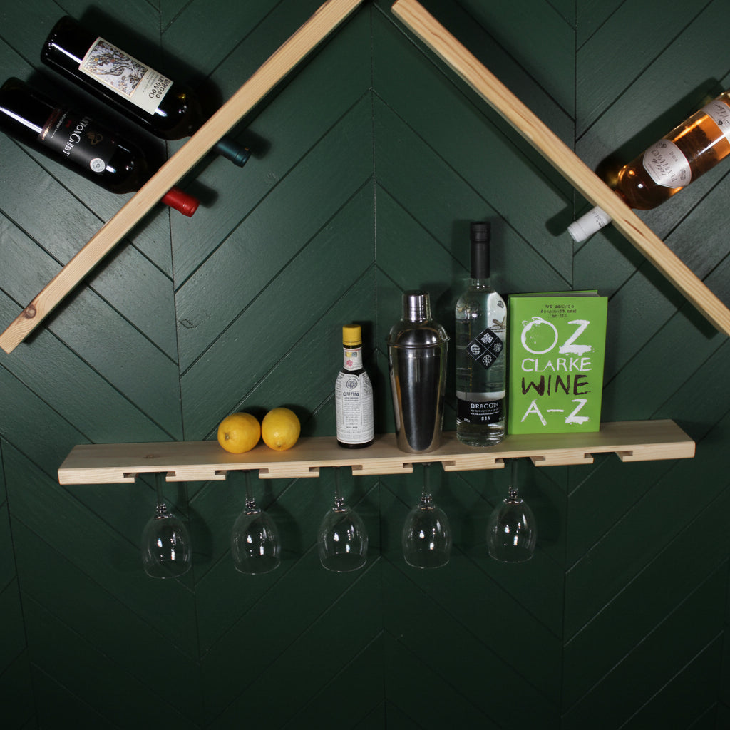A house-shaped wooden wall-mounted wine rack with a lower shelf holding wine glasses, lemons, bottles, and a green 'Oz Wine A-Z' box, mounted on a white wall.