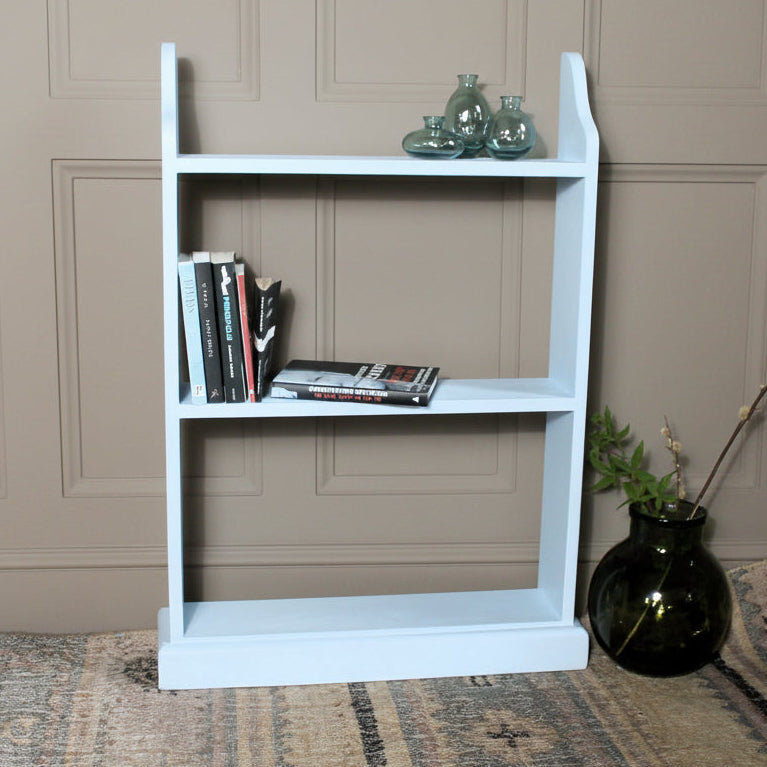 A light blue three-tier wooden bookshelf with decorative items on its shelves, set against a rustic gray brick wall on a patterned rug.