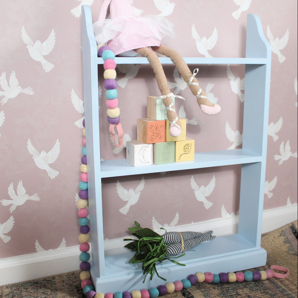 A light blue three-tier wooden shelf styled with a pink ballerina doll, pastel blocks, a beaded garland, and a small bouquet of green leaves, set against a pink wallpaper.